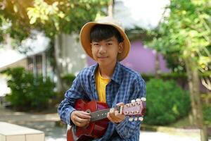 ett asiatisk pojke spelar de ukulele under hans fri tid i de skola gård under de träd. mjuk och selektiv fokus. foto