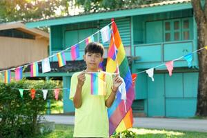 asiatisk pojke innehar en regnbåge flagga i främre av en hus dekorerad med regnbåge flaggor under stolthet månad till visa HBTQ stolthet och identitet. mjuk och selektiv fokus. foto