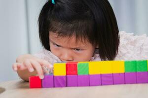 närbild söt asiatisk flicka spelar med färgrik trä- kub. barn spänt syftar till på placering av trä- block. fri tid och inlärning genom spela. unge åldrig 4 år gammal. foto