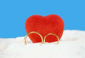 röd hjärta med bröllop guld ringar på sand av strand Bakom hav. hjärtans dag, bröllop, smekmånad, Semester. kopia Plats foto