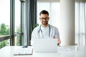 läkare med bärbar dator utför ett uppkopplad video ring upp och samtal till patient. manlig läkare arbetssätt på kontor skrivbord, kontor interiör på bakgrund foto