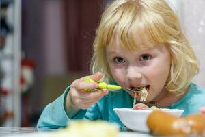en liten flicka har frukost på Hem spaghetti med korvar. liten blond flicka äter middag med gaffel på tabell foto