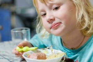 en liten flicka har frukost på Hem spaghetti med korvar. liten blond flicka äter middag med gaffel på tabell foto