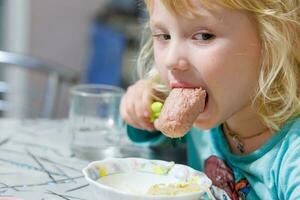 en liten flicka har frukost på Hem spaghetti med korvar. liten blond flicka äter middag med gaffel på tabell foto