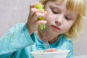 en liten flicka har frukost på Hem spaghetti med korvar. liten blond flicka äter middag med gaffel på tabell foto