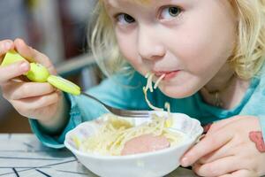 en liten flicka har frukost på Hem spaghetti med korvar. liten blond flicka äter middag med gaffel på tabell foto
