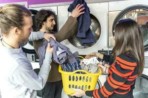 ung grupp av tusenåriga vänner är sätta kläder i de tvättning maskin foto