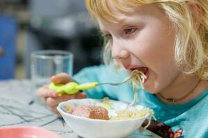 en liten flicka har frukost på Hem spaghetti med korvar. liten blond flicka äter middag med gaffel på tabell foto