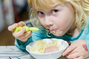 en liten flicka har frukost på Hem spaghetti med korvar. liten blond flicka äter middag med gaffel på tabell foto