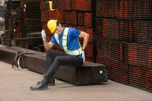 porträtt av en manlig arbetstagare bär en säkerhet väst och hjälm Sammanträde på en stål lastpall på grund av till tillbaka smärta från arbetssätt i en fabrik lyft tung saker. foto