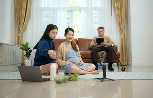 en gravid asiatisk kvinna sitter på de golv med henne yngre syster. leva social media utsända försäljning gravid mödrar Produkter och måltider, där är en far i de bakgrund avkopplande på en soffa. foto