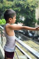 liten unge spelar i sommar regn i hus balkong, indisk smart pojke spelar med regn droppar under monsun regnig säsong, unge spelar i regn foto