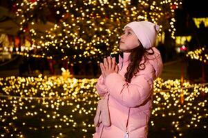 söt unge flicka med händer kupade, framställning önskar för jul, mot en gata upplyst förbi jul lampor på en rättvis foto