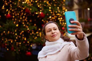 skön mång etnisk kvinna framställning selfie på henne smartphone, stående mot upplyst jul träd medan tivoli foto