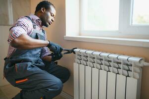 afro reparatör i overall använder sig av verktyg medan montera eller reparation uppvärmning radiator foto