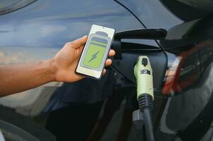 man innehav smartphone medan laddning bil på elektrisk fordon laddning station, närbild. foto