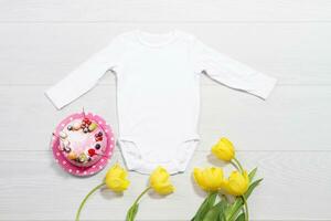 vit attrapp bebis nyfödd skjorta. trä- bakgrund, gul tulpaner, Lycklig födelsedag rosa kaka. tom mall overall kroppsdräkt främre topp se. bebis Kläder. bukett av blommor foto