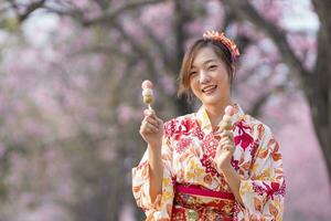 japansk kvinna i traditionell kimono klänning innehav ljuv hanami dango efterrätt medan gående i de parkera på körsbär blomma träd under vår sakura festival foto