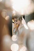 blond brud poser i en morgonrock och tiara, bröllop porträtt. reflektioner och reflektioner av glas. bröllop Foto i en ljus nyckel