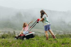 en kvinna i stövlar med henne barn i de form av en spel klipper de gräs med en gräsklippare i de trädgård mot de bakgrund av bergen och dimma, trädgård verktyg begrepp foto