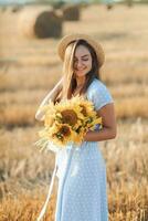 porträtt av en ung flicka. en flicka i en blå klänning och hatt, innehav en bukett av solrosor, mot en bakgrund av hö balar. lång hetero hår. trevlig Färg. sommar foto