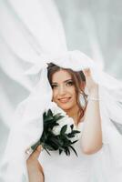 lockigt brunett brud i en vit klänning, täckt med en slöja, poser för de kamera med en bukett av rosor. porträtt av de brud. skön smink och hår. bröllop i natur foto