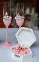 guld bröllop ringar på en dekorativ vit låda med rosa blommor liggande på en vit tabell Nästa till rosa bröllop vin glasögon, närbild. Smycken begrepp foto