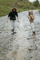 två Lycklig liten flickor av europeisk utseende spelar i vattenpölar under regn i sommar. barn är spelar i de regn. barn spelar i natur utomhus. de flicka åtnjuter de regn. foto