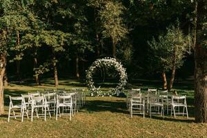 bröllop ceremoni i de skog. de väg till de runda båge med lövverk, gröna, grönska, och blommor. rustik dekor. trä- stolar i de bakgård bankett område. säten för gäster. foto