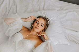 en skön lockigt brun håriga brud i en vit klänning poser för en fotograf medan liggande på en säng i en skön klänning med ärmar. bröllop fotografi, närbild porträtt, chic frisyr. foto