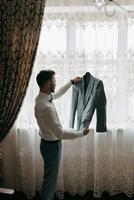 en man i en vit skjorta innehar hans grå jacka, mot de bakgrund av en fönster. sida se. en eleganta Kolla på. herr- stil. mode. företag foto