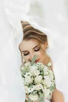 lockigt blond brud i en vit klänning, täckt med en slöja, poser för de kamera med en bukett av rosor. porträtt av de brud. skön smink och hår. bröllop i natur foto