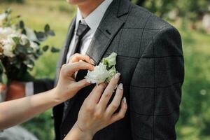 de brud är innehav en boutonniere. beskurna Foto av brudens händer justeras boutonniere på brudgummens jacka på bröllop ceremoni. de brudgum är innehav en bukett. franska manikyr. bröllop detaljer