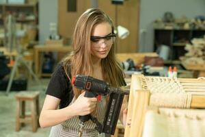 kvinna snickare bär skyddande säkerhet glasögon och använder sig av elektrisk arbete på en trä. konstnär eller möbel designer arbetssätt på en produkt aning i en verkstad. foto
