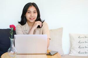 Lycklig asiatisk kvinna Sammanträde på soffa använder sig av bärbar dator och smartphone ser kamera med leende bär tillfällig kläder modern foto