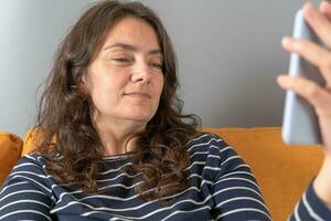 attraktiv medelålders kvinna talande på smartphone genom video ring upp medan Sammanträde på soffa foto