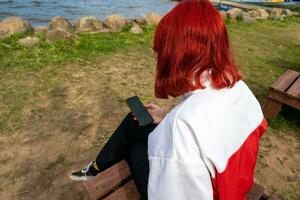 flicka med röd hår, generation z, sitter på de Strand av en tyst sjö och användningar en smartphone foto