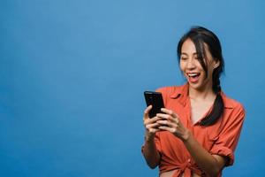 ung asiatisk dam som använder telefon med positivt uttryck, ler brett, klädd i vardagskläder som känner lycka och står isolerad på blå bakgrund. glad förtjusande glad kvinna jublar över framgång.