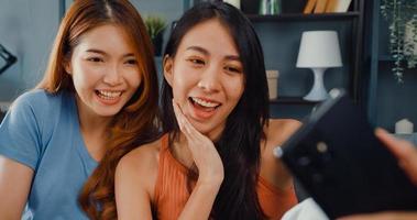 tonåring asiatiska kvinnor som känner sig glada leende slappna av använder smartphone videosamtal i vardagsrummet hemma. glada rumskamrat damer videokonferens med vän och familj, livsstil kvinna hemma koncept. foto