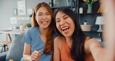 tonåring asiatiska kvinnor känner sig lyckliga leende selfie och tittar på kameran medan de kopplar av i vardagsrummet hemma. glada rumskamrat damer videosamtal med vän och familj, livsstil kvinna hemma koncept. foto