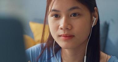 ung asiatisk tjej tonåring med slitage hörlurar använda bärbar dator lära sig online skriva föreläsning anteckningsbok i vardagsrummet hemma. isolera utbildning online e-learning coronavirus-pandemikoncept. foto