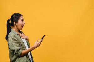 förvånad ung asiatisk dam med mobiltelefon med positivt uttryck, ler brett, klädd i vardagskläder och står isolerad på gul bakgrund. glad förtjusande glad kvinna jublar över framgång.