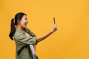 le bedårande asiatisk kvinna som gör selfie foto på smart telefon med positivt uttryck i vardagskläder och stativ isolerad på gul bakgrund. glad förtjusande glad kvinna jublar över framgång.