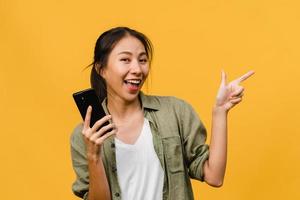 ung asiatisk dam som använder mobiltelefon med glada uttryck, visar något fantastiskt på tomt utrymme i vardagsduk och tittar på kameran isolerad över gul bakgrund. ansiktsuttryck koncept. foto