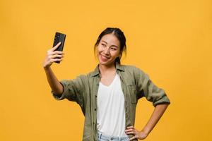 le bedårande asiatisk kvinna som gör selfie foto på smart telefon med positivt uttryck i vardagskläder och stativ isolerad på gul bakgrund. glad förtjusande glad kvinna jublar över framgång.