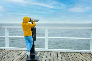 en pojke i en gul jacka och jeans, stående på de pir, ser genom kikare på de baltic hav. kall väder foto