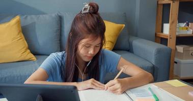 ung asiatisk tjej tonåring med fritidsdräkt hörlurar använda digital surfplatta lära sig online skriva föreläsning anteckningsbok i vardagsrummet hemma. isolera utbildning online e-learning coronavirus-pandemikoncept. foto