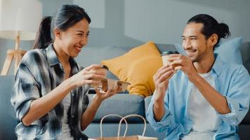 lyckliga asiatiska unga attraktiva par man och kvinna sitter vid nytt hem dricka kaffe koppla av och prata leende med kartongförpackning förvaring för att flytta i nytt hus. unga gifta asiatiska flytta hem koncept.