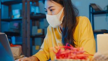 ung asiatisk affärskvinna bära ansiktsmask kontrollera inköpsorder och svara klient inkorg chatt på bärbar dator på hemmakontor på natten. småföretagare, onlinemarknadsleverans, livsstil frilansande koncept.