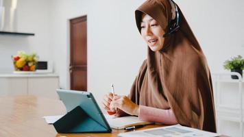 asien muslimsk dam bär hörlurar med digital surfplatta prata med kollegor om försäljningsrapport i konferensvideosamtal medan du arbetar hemma i köket. social distansering, karantän för corona.